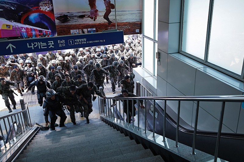 Grupa umundurowanych policjantów w hełmach i tarczach biegnie po schodach w metrze lub na stacji kolejowej, pod niebieskimi znakami i dużymi plakatami, przez okna wpadają promienie słońca.