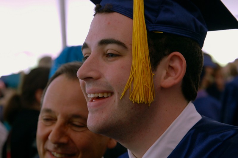 Młody mężczyzna w niebieskiej czapce i todze uśmiecha się radośnie, stojąc obok starszego mężczyzny na zatłoczonej ceremonii ukończenia szkoły. Żółty frędzel zwisa z czapki.