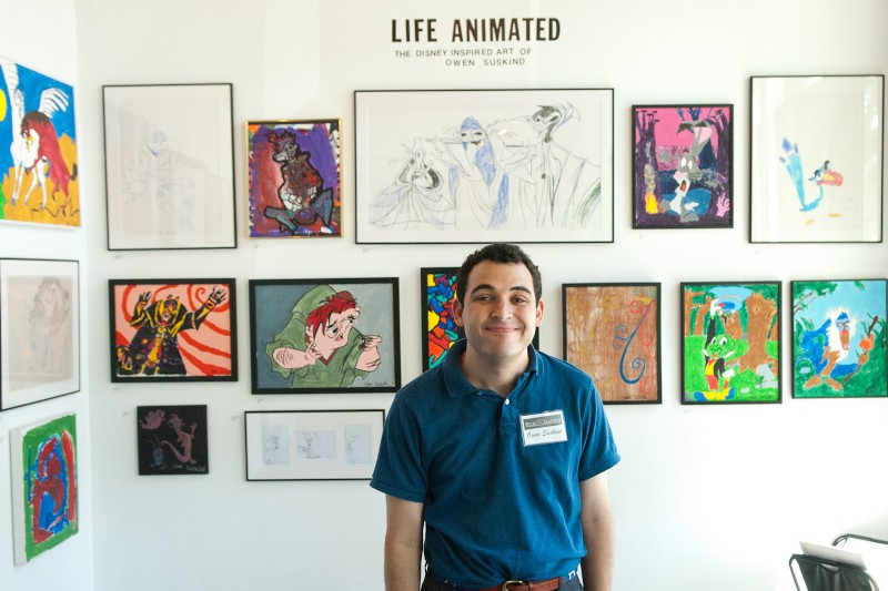 Mężczyzna w niebieskiej koszuli stoi uśmiechnięty przed ścianą wypełnioną kolorowymi oprawionymi dziełami sztuki. Nad nim tekst brzmi LIFE ANIMATED, The Disney Inspired Art of Owen Suskind.