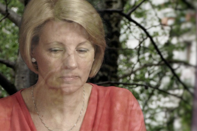 Kobieta o krótkich blond włosach i czerwonej bluzce spogląda zamyślona w dół, na co patrzy przez okno, w którym gałęzie drzew odbijają się w szybie.