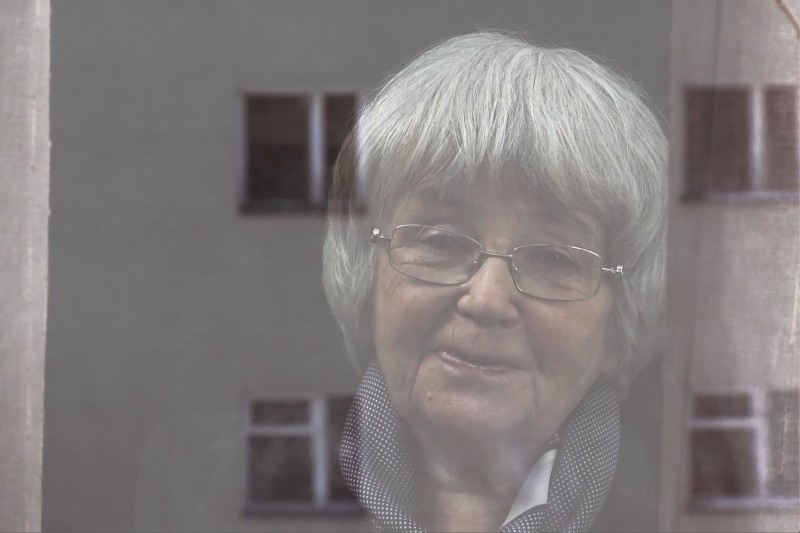 Starsza kobieta z siwymi włosami i okularami patrzy przez okno, jej twarz częściowo odbija się w szkle. Ma na sobie ciemną kurtkę i szalik, a w tle widać rozmazane okna budynku.