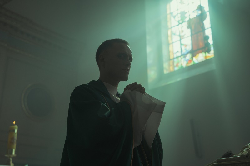 Osoba ubrana w szatę liturgiczną stoi wewnątrz słabo oświetlonego kościoła, ze złożonymi rękami do modlitwy, a w tle widać światło wpadające przez witraż w oknie.