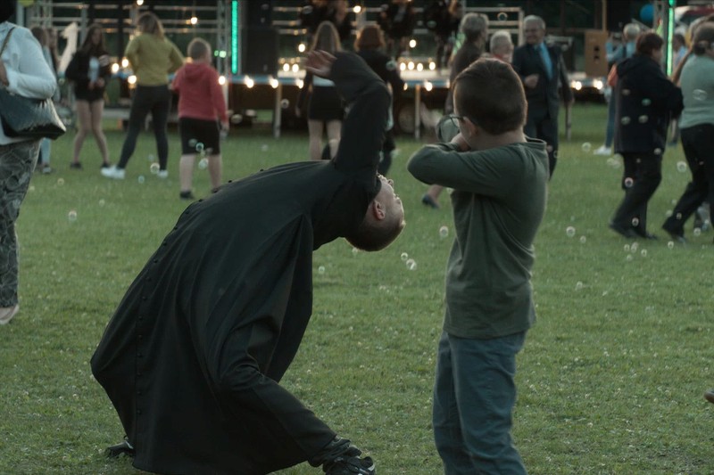 Osoba w długim, czarnym płaszczu dramatycznie odchyla się do tyłu obok dziecka, które zakrywa twarz na trawiastym terenie podczas imprezy plenerowej, w tle widać ludzi i scenę.