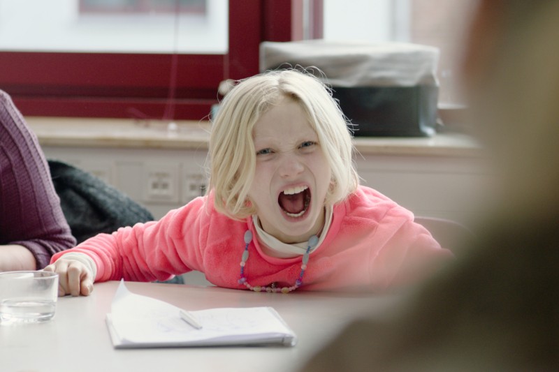 Małe dziecko o blond włosach, ubrane w różową bluzę i kolorowy naszyjnik, siedzi przy stole z papierem i szklanką, pochyla się do przodu i krzyczy z szeroko otwartymi ustami.