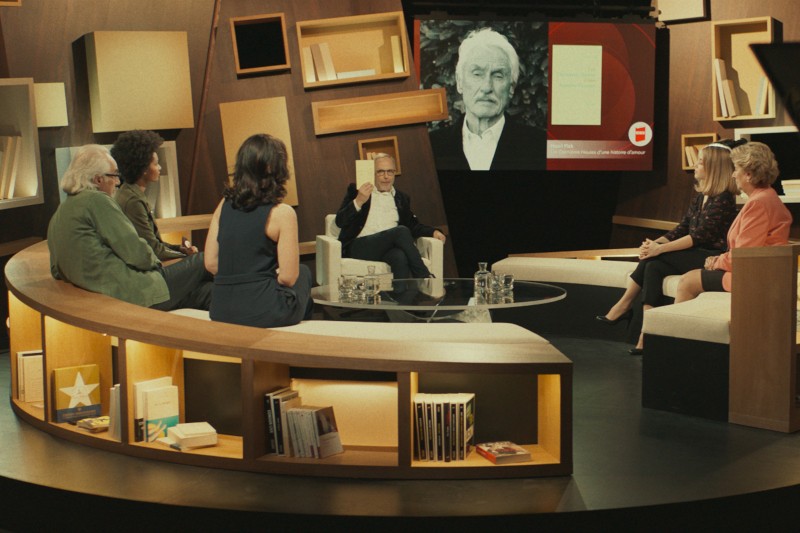 Grupa ludzi siedzi w półkolu na nowoczesnym planie telewizyjnym, angażując się w dyskusję. Książki są wystawione na półkach, a duży ekran w tle pokazuje portret starszego mężczyzny w skali szarości.