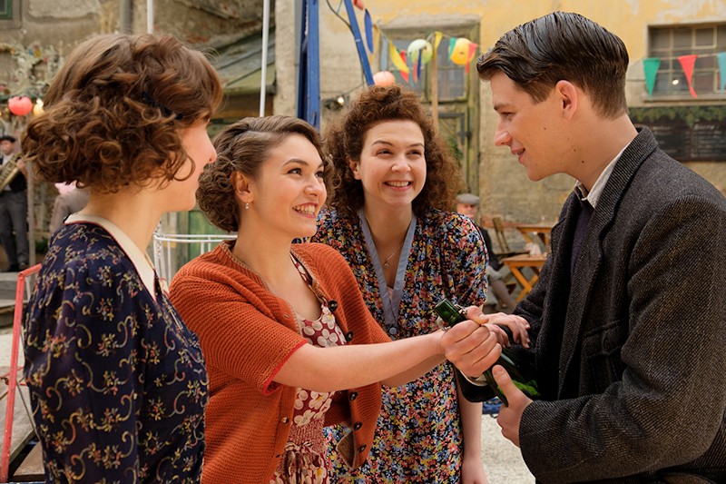 Trzy uśmiechnięte kobiety w sukienkach vintage rozmawiają radośnie z młodym mężczyzną trzymającym butelkę, na zewnątrz, w otoczeniu kolorowych flag i lampionów. Scena ma nostalgiczny klimat połowy XX wieku.