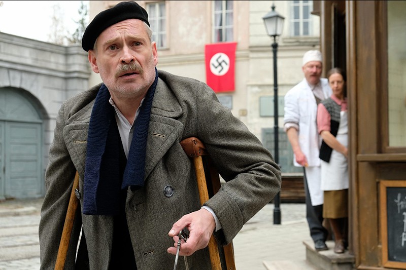 Mężczyzna o kulach i w kapeluszu wygląda na zaniepokojonego na tle ulicy, na której powiewa flaga nazistowska; dwie osoby w białych fartuchach stoją przy budynku i obserwują go.