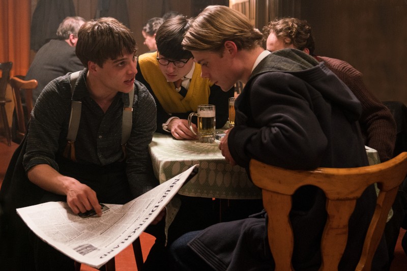 Trzech młodych mężczyzn siedzi blisko siebie przy stole w słabo oświetlonym pokoju, uważnie studiując gazetę. Jeden wskazuje na gazetę, podczas gdy drugi trzyma kufel piwa. Czwarta osoba jest rozmazana w tle.