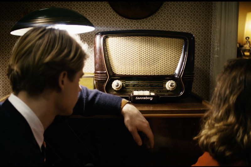 Dwie osoby siedzą blisko siebie, naprzeciwko staromodnego radia stołowego na drewnianej powierzchni, w ciepło oświetlonym pokoju z wzorzystą tapetą. Radio jest oświetlone lampą nad nim.