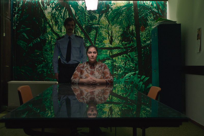 Kobieta siedzi przy odbijającym stole przed muralem z gęstej zieleni liści, a za nią stoi mężczyzna w mundurze policyjnym. W pomieszczeniu jest słabo oświetlone i znajdują się brązowe krzesła.