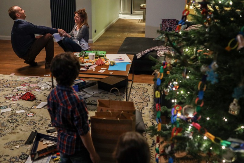 Mężczyzna żartobliwie trzyma kobietę za ręce, gdy siedzi uśmiechnięta w przytulnym salonie. Dwoje dzieci stoi w pobliżu, a udekorowana choinka i porozrzucane gry i rękodzieło pokrywają podłogę.