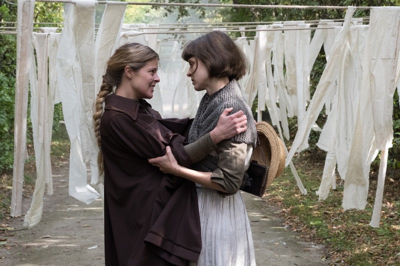 Dwie kobiety stoją blisko siebie, patrząc sobie w oczy, otoczone białymi szmatami wiszącymi na sznurku na zewnątrz w zalesionym terenie. Jedna kobieta trzyma słomkowy kapelusz i delikatnie chwyta ramiona drugiej.