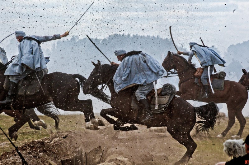 Mężczyźni w historycznych mundurach wojskowych jadą konno przez pole bitwy, z podniesionymi mieczami, a w powietrzu unosi się kurz i gruz, co tworzy dramatyczną scenę akcji.