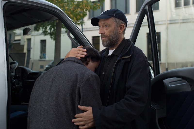 Mężczyzna w czarnej kurtce i czapce delikatnie przytula i pociesza inną osobę, która opiera głowę na jego piersi, przy otwartych drzwiach samochodu na miejskiej ulicy.