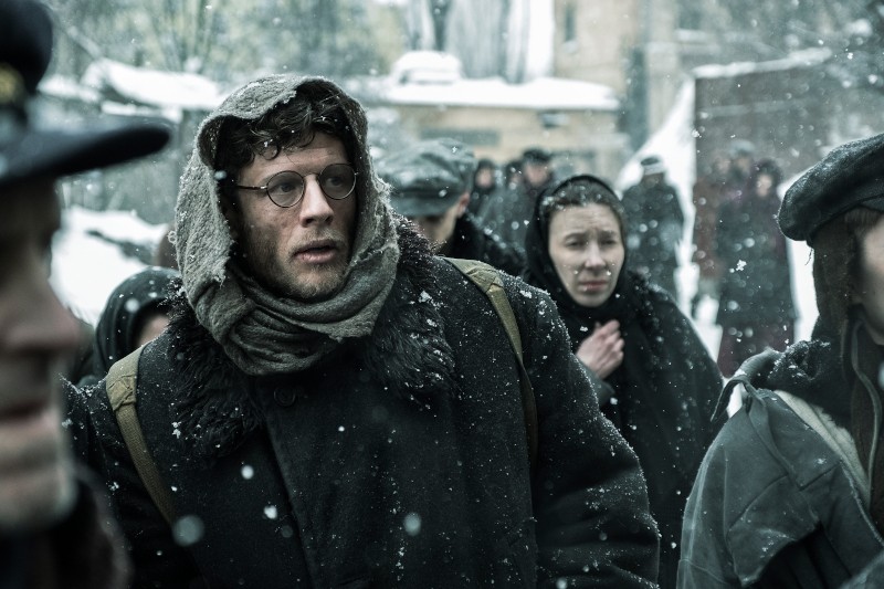 Grupa ludzi w zimowych ubraniach idzie przez padający śnieg; jeden mężczyzna na pierwszym planie nosi okulary, gruby szalik i kaptur, patrzy przed siebie z ponurym wyrazem twarzy, podczas gdy inni idą za nim.