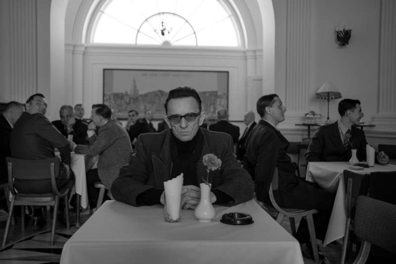 Mężczyzna w ciemnych okularach siedzi sam przy stoliku kawiarnianym z wazonem z kwiatami, otoczony przez innych mężczyzn w garniturach, którzy rozmawiają. Otoczenie jest eleganckie, z dużymi oknami i klasycznym wystrojem w czerni i bieli.