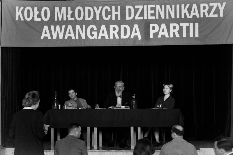 Czarno-białe zdjęcie przedstawia trzy osoby siedzące przy stole na scenie pod polskim transparentem. Przed nimi stoi publiczność i kobieta, prawdopodobnie na spotkaniu lub konferencji.
