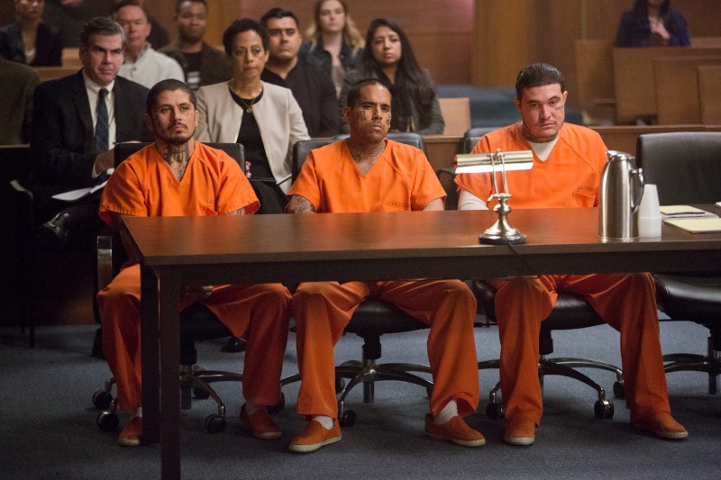 Trzech mężczyzn w pomarańczowych mundurach więziennych siedzi obok siebie przy stole w sali sądowej, zwróconych twarzą do przodu. Ludzie siedzą na galerii za nimi, obserwując przebieg rozprawy. Na stole leży metalowy dzbanek i notatniki prawnicze.