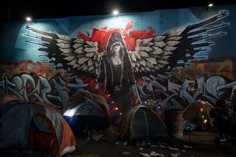 Na niebieskiej ścianie, nad kilkoma namiotami i porozrzucanymi rzeczami, namalowano mural przedstawiający zakapturzoną postać z piórami na skrzydłach i bronią. Sceneria nocnego obozowiska w mieście.