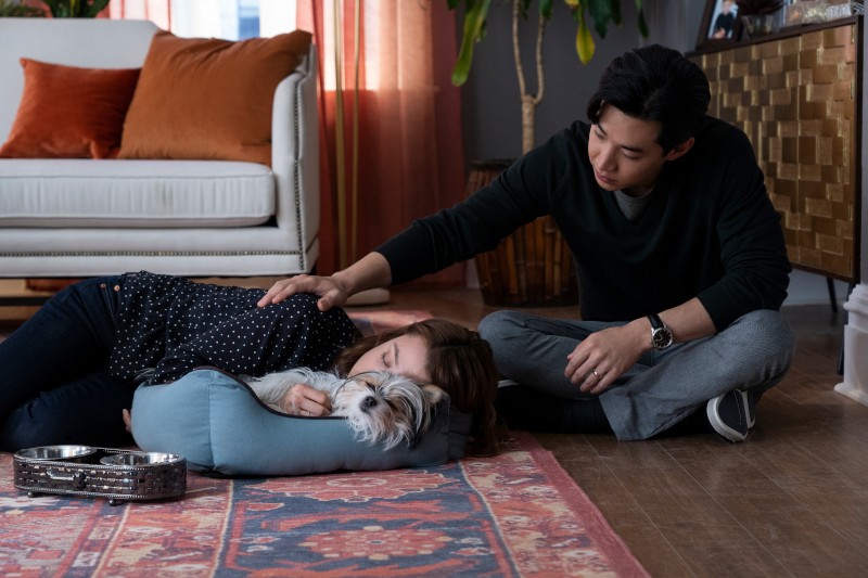 Kobieta leży zwinięta w legowisku obok małego psa, podczas gdy mężczyzna siedzi obok niej na podłodze i delikatnie kładzie rękę na jej plecach w przytulnym salonie.