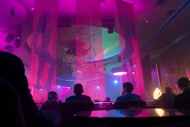 Ludzie siedzący w słabo oświetlonym klubie nocnym z różowym oświetleniem, obserwujący sylwetki tancerzy za cienkimi firankami. Atmosfera jest żywa dzięki reflektorom, żyrandolom i odbijającym światło stołom.