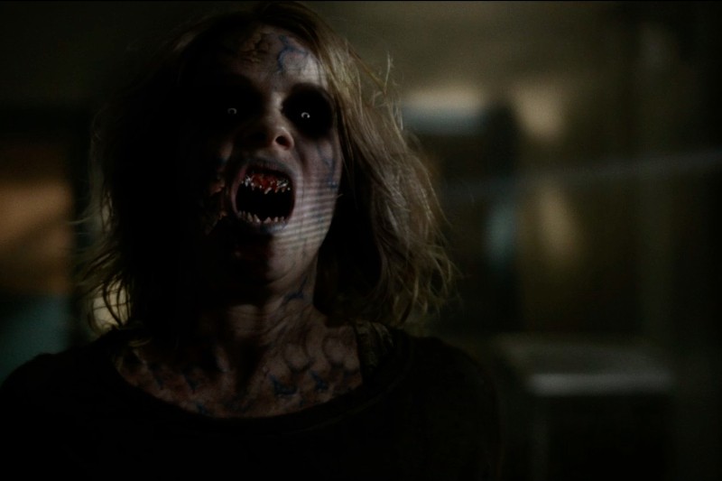 Osoba o bladej skórze, ciemnych oczach, ostrych, spiczastych zębach i zniekształconych ustach, oświetlona w słabym świetle, wydaje się potworna i przerażająca.