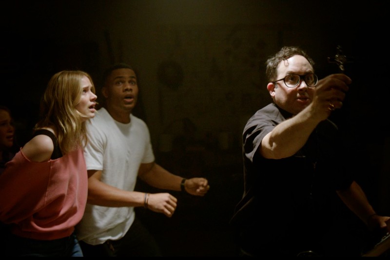 Trzy osoby, w tym dwie przestraszone, stoją za mężczyzną w okularach trzymającym krucyfiks w ciemnym, zadymionym pomieszczeniu, jakby odpędzały coś niewidzialnego.