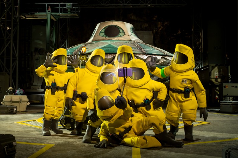 Sześć osób w żółtych kombinezonach ochronnych pozuje do grupowego selfie przed futurystycznym statkiem kosmicznym w hangarze high-tech. Scena wydaje się zabawna pomimo poważnego otoczenia.