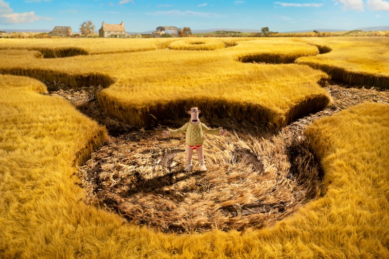 Mężczyzna w jasnej kurtce stoi z wyciągniętymi rękami pośrodku kręgu zbożowego na złotym polu pszenicy, na tle którego widać dom i drzewa pod błękitnym niebem.