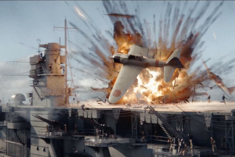 Samolot myśliwski z czasów II wojny światowej rozbija się na pokładzie lotniskowca, powodując potężną eksplozję, w której pojawiają się płomienie i odłamki, a ludzie uciekają, szukając schronienia.