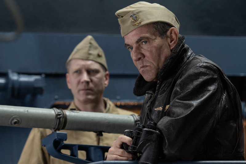 Dwóch mężczyzn w mundurach wojskowych stoi na czymś, co wygląda jak statek lub łódź podwodna. Mężczyzna z przodu nosi skórzaną kurtkę i czapkę, trzyma lornetkę i wygląda poważnie; mężczyzna z tyłu nosi mundur khaki i czapkę, patrzy przed siebie.