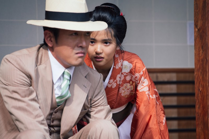 Mężczyzna w beżowym garniturze i kapeluszu siedzi z przodu, podczas gdy kobieta w tradycyjnym pomarańczowym kimonie kuca za nim, patrząc uważnie przed siebie. Wyglądają, jakby byli zaangażowani w poważną rozmowę.