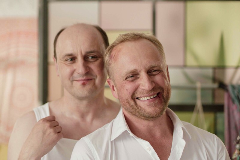 Dwóch mężczyzn stoi blisko siebie w pomieszczeniu. Mężczyzna z przodu, ubrany w białą koszulę, szeroko się uśmiecha, podczas gdy mężczyzna za nim, w bezrękawnikowej koszulce, ma łagodny wyraz twarzy. Tło zdobią panele w pastelowych kolorach.