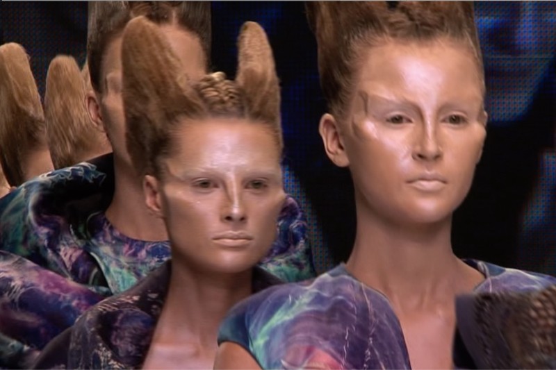 Modelki z wyrzeźbionymi, wysoko upiętymi włosami i futurystycznym makijażem chodzą po wybiegu, ubrane w kolorowe, wzorzyste ubrania ze strukturalnymi ramionami. Tło jest ciemne, co przyciąga uwagę do ich awangardowego wyglądu.