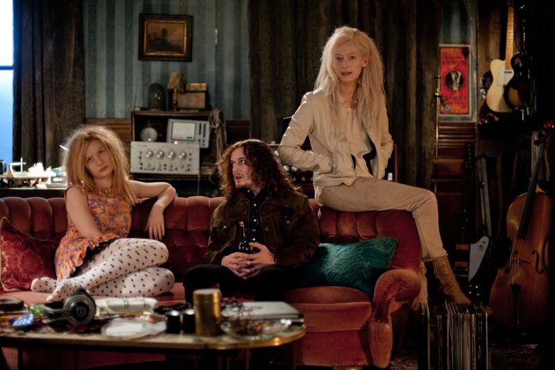 Trzy osoby siedzą na czerwonej sofie w słabo oświetlonym, zagraconym pokoju z instrumentami muzycznymi i starą elektroniką. Dwie mają długie włosy i swobodne ubrania; jedna siedzi na oparciu sofy, ubrana w lekką kurtkę i buty.