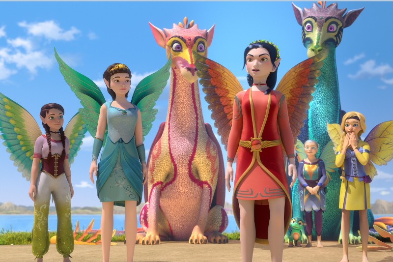 Pięć animowanych postaci wróżek z kolorowymi skrzydłami stoi na piasku obok dwóch dużych, przyjaznych smoków i małej żaby, z jasnoniebieskim niebem i wodą w tle. Scena wydaje się radosna i magiczna.