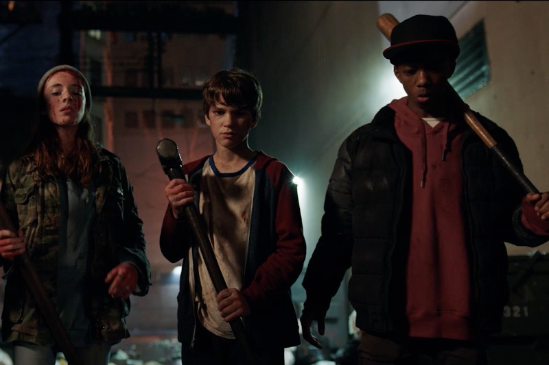 Trójka dzieci stoi blisko siebie w słabo oświetlonej alejce, każde trzyma broń — kij lub fajkę — wyglądając poważnie i zdeterminowanie. Noszą luźne, wytrzymałe ubrania i mają smugi na twarzach, sugerujące niedawne działania lub konflikty.