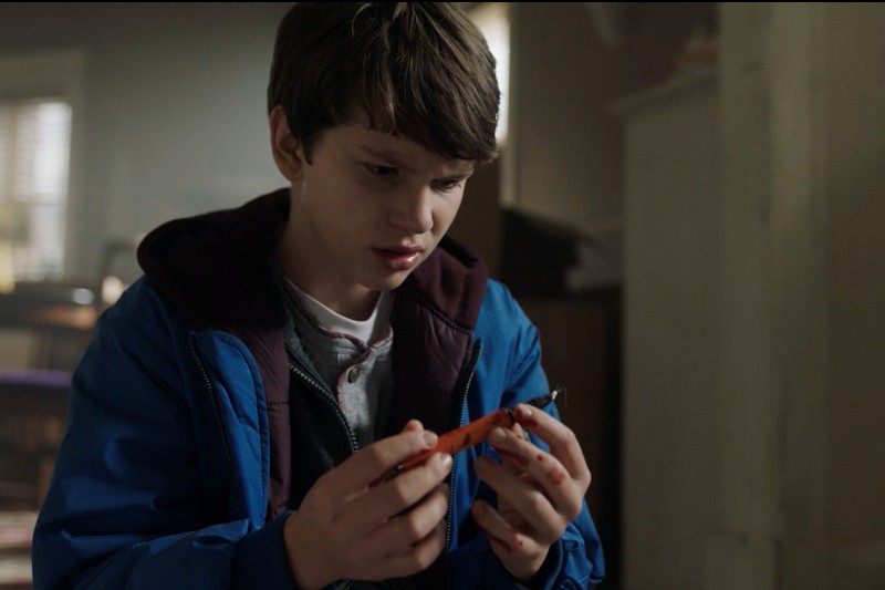 Młody chłopiec o brązowych włosach, ubrany w niebieską kurtkę, siedzi w środku i uważnie patrzy na pomarańczowy przedmiot, który trzyma w rękach. Pokój za nim jest delikatnie oświetlony i wydaje się zagracony.