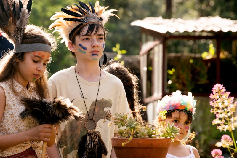 Trójka dzieci w nakryciach głowy z piór i pomalowanych twarzach stoi na zewnątrz w słońcu, otoczona roślinami. Dwoje trzyma wachlarze z piór i wydaje się, że biorą udział w zabawie lub aktywności kulturalnej.