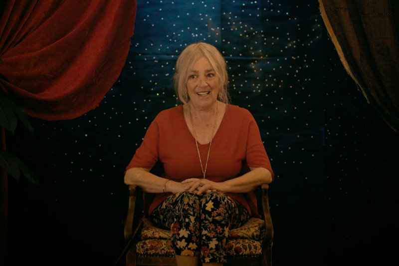 Starsza kobieta z siwymi włosami, ubrana w czerwoną bluzkę i kwieciste spodnie, siedzi uśmiechnięta na wzorzystym krześle na tle rozgwieżdżonego nieba, otoczonego czerwonymi i brązowymi zasłonami.