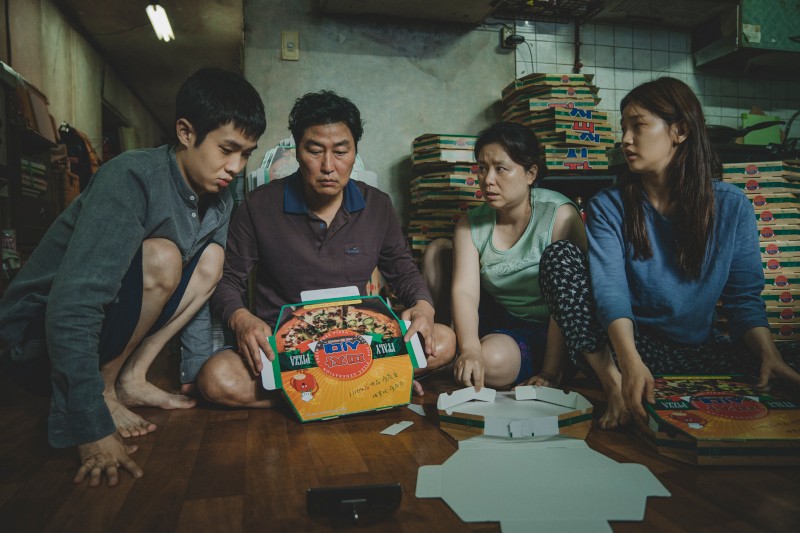 Cztery osoby siedzą na podłodze w zagraconym pokoju, składając pudełka po pizzy, wyglądając poważnie i skupione. Pudełka po pizzy są ułożone wysoko w tle, a pokój wydaje się ciasny i słabo oświetlony.