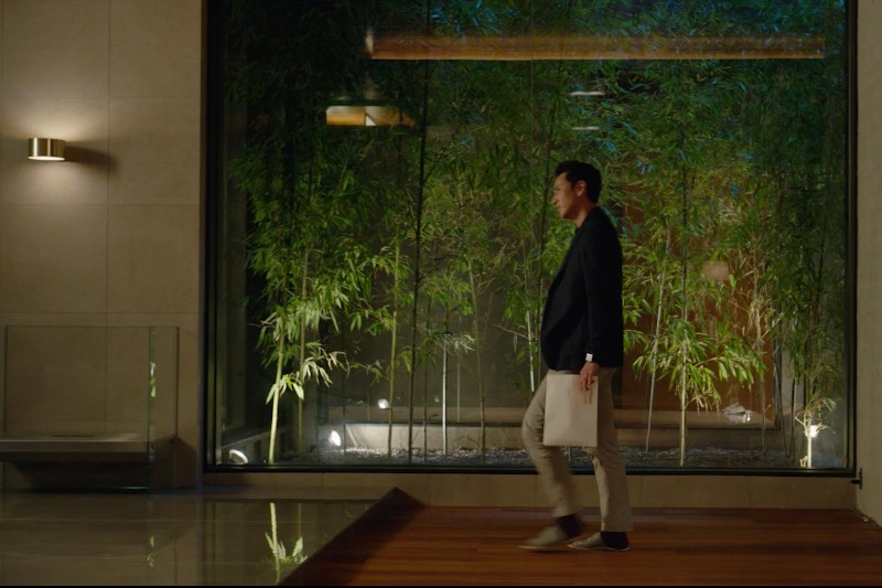 Mężczyzna w marynarce i jasnych spodniach wchodzi do środka, trzymając teczkę. Za nim duże okna odsłaniają wysokie rośliny bambusa oświetlone delikatnym światłem, tworząc nowoczesną i spokojną atmosferę.