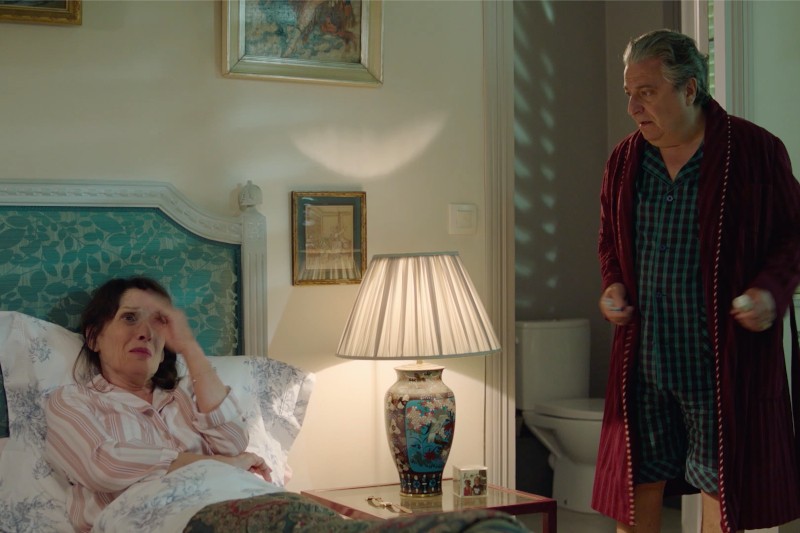 Kobieta w piżamie siedzi na łóżku, wyglądając na zmartwioną, z ręką na czole, podczas gdy starszy mężczyzna w szlafroku i piżamie stoi w pobliżu, wyglądając na zaniepokojonego. W pokoju widać lampę i oprawione dzieło sztuki.