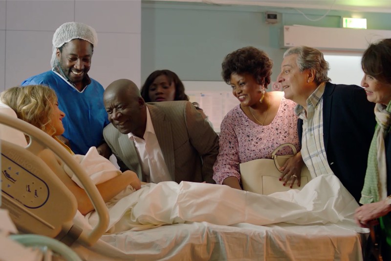Grupa ludzi zbiera się wokół kobiety leżącej na szpitalnym łóżku, uśmiechając się i patrząc na nią i noworodka. Scena wydaje się radosna i pełna świętowania w szpitalnej sali.