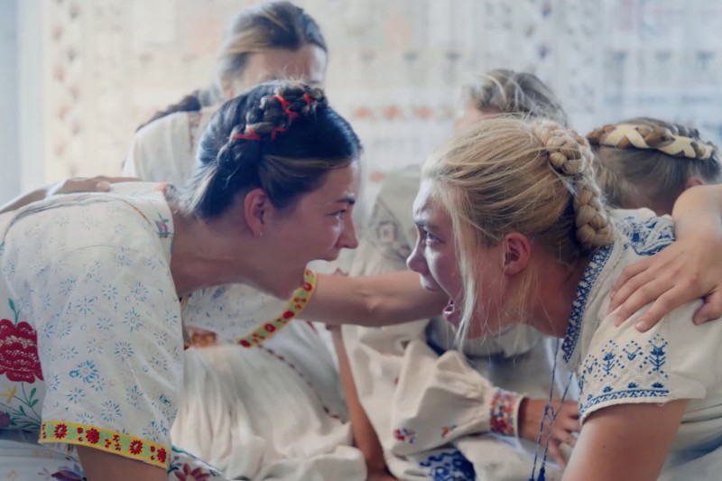 Dwie kobiety ubrane w tradycyjne haftowane suknie siedzą naprzeciwko siebie, namiętnie płacząc i krzycząc, otoczone innymi w podobnych strojach. Trzymają się za ręce w emocjonalnym, intensywnym momencie.