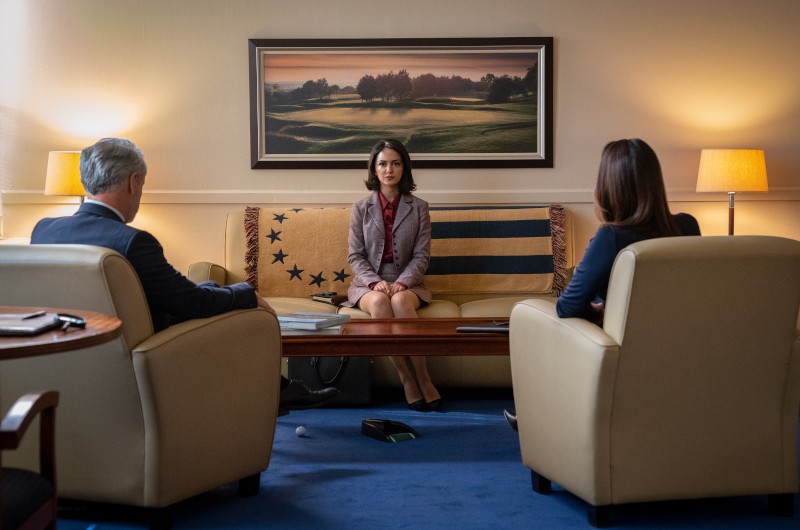 Kobieta siedzi na kanapie między dwoma osobami w fotelach naprzeciwko niej, w formalnym biurze z obrazem krajobrazu, lampami i flagą USA narzuconą za nią. Scena sugeruje poważną rozmowę lub spotkanie.