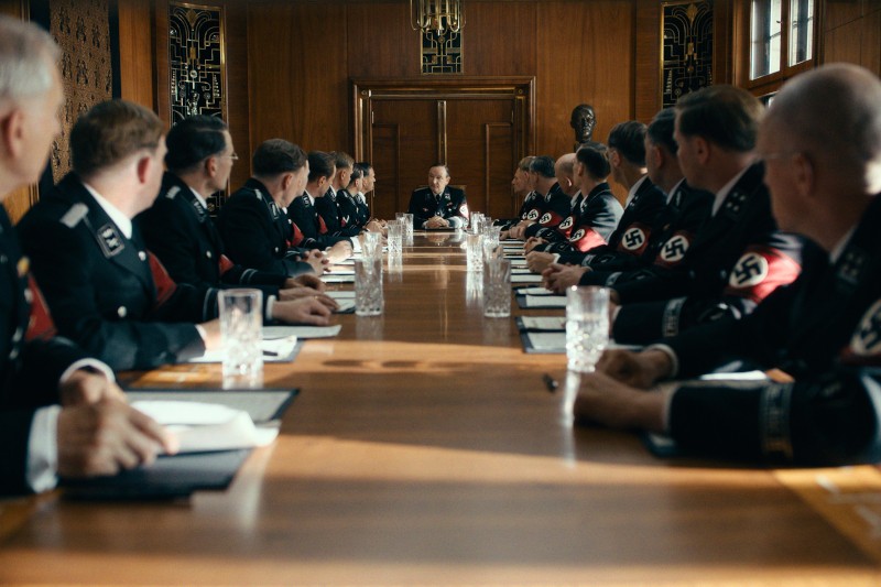 Grupa mężczyzn w mundurach nazistowskich SS siedzi wokół długiego drewnianego stołu konferencyjnego w formalnej sali konferencyjnej. Mężczyzna siedzący na czele stołu wydaje się przewodzić dyskusji.