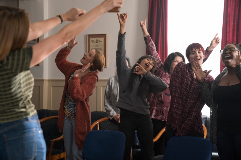 Grupa kobiet stoi w pokoju, śmiejąc się i tańcząc z uniesionymi ramionami, wyglądając na radosne i energiczne. Na pierwszym planie widoczne są puste niebieskie krzesła.