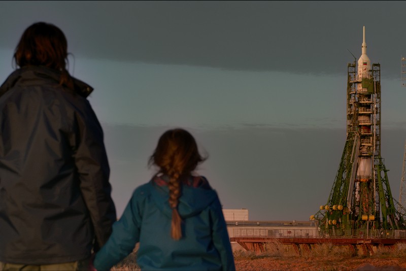Dwie osoby, dorosły i dziecko, stoją na pierwszym planie, trzymając się za ręce i obserwując dużą rakietę na platformie startowej o zachodzie słońca, na tle dramatycznego nieba.