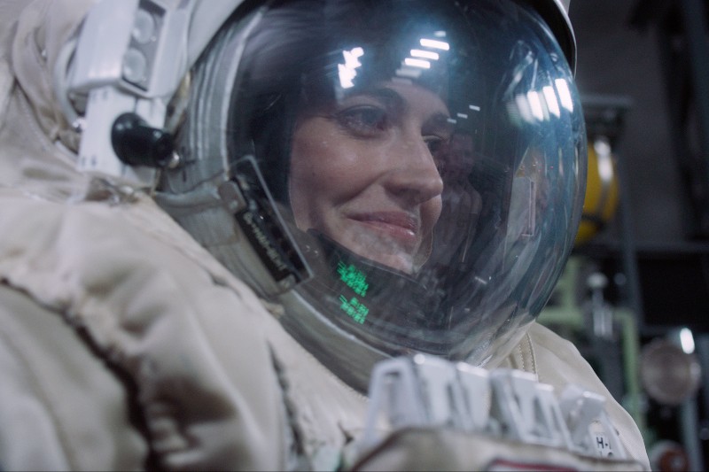 Uśmiechnięty astronauta ubrany w hełm i skafander odbija się w szkle hełmu, w tle widać światła i sprzęt.